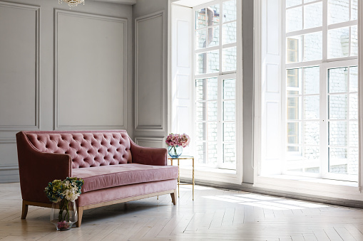 White room with pink sofa-bed, flowers in glass jug and mirror near the window. Classic interior design