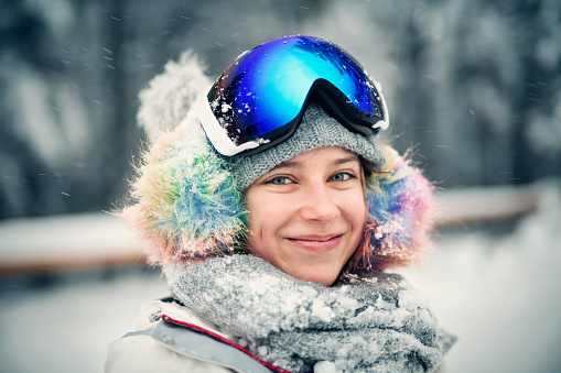 Happy little girl in ski outfit smiling at the camera. The girl is wearing ski goggles. It's snowing on a winter day.\nNikon D850