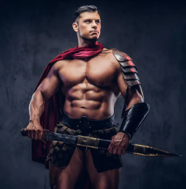 Brutal ancient Greek warrior with a muscular body in battle equipment posing on a dark background.