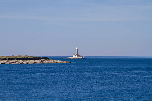 This serene image captures a soft summer sunset along the rocky coast of Hvar Island, Croatia. A small, white lighthouse stands alone on a stone pier, facing the gentle Adriatic Sea. The pastel-toned sky, transitioning from warm oranges to soft blues, reflects the peacefulness of a Mediterranean evening. The scene is perfect for themes of solitude, nature, coastal travel, and calm reflections.