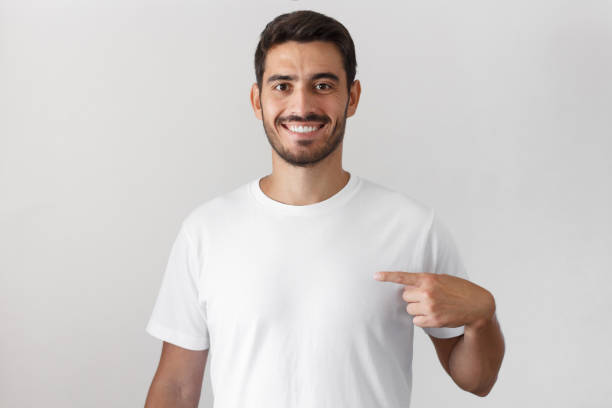 jovem bonito apontando com o dedo indicador na t-shirt branca em branco com espaço vazio para a sua publicidade texto ou imagem, em pé isolado em fundo cinza - aparência - fotografias e filmes do acervo