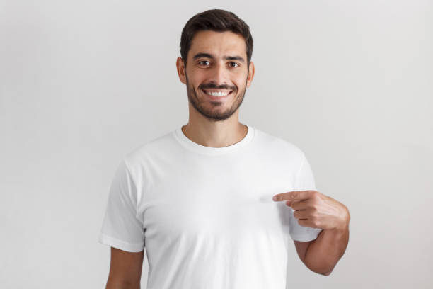 istock Horizontal shot of young handsome caucasian man isolated on gray background, smiling while pointing with index finger to white blank t-shirt, copyspace for advertising 1029759356