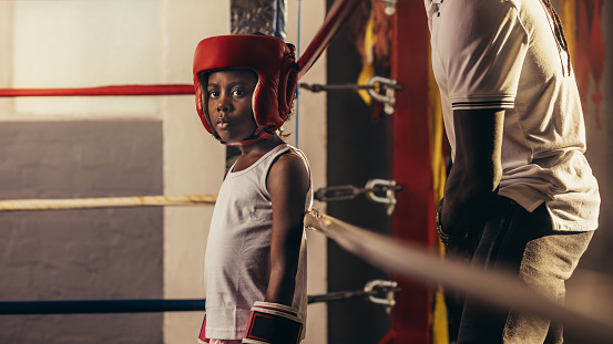 Kid wearing boxing gloves and headgear standing in a corner of a boxing ring. Small boy standing inside a boxing ring with his coach behind him.