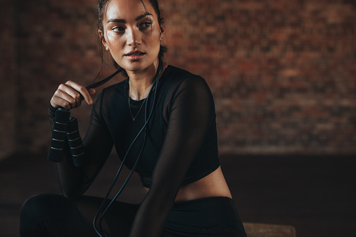Fit woman in sportswear sitting with skipping rope and looking away. Female athlete taking break after training session at gym.