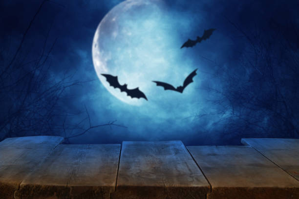 istock Halloween holiday concept. Empty rustic table in front of scary and misty night sky with black bats and full moon background. Ready for product display montage. 1029791220