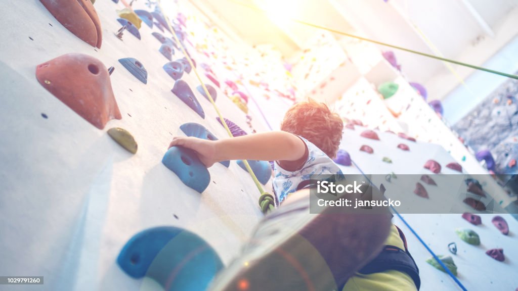 young boy is climbing an indoor climbing wall closeup of young boy hanging on a rope leading to the top of an artificial climbing wall, personal development and career growth or challenge yourself concept Climbing Stock Photo