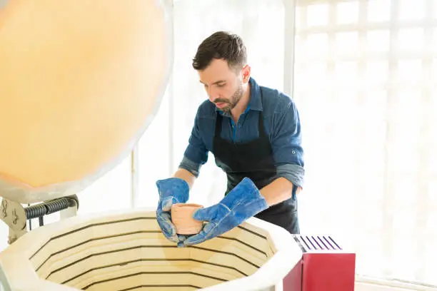 Mid adult man wearing gloves while placing clay mug into kiln at pottery studio