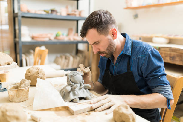 Owner Making Clay Sculpture Of Doll In Workshop Confident male owner making clay sculpture of doll at workbench in workshop pottery making stock pictures, royalty-free photos & images