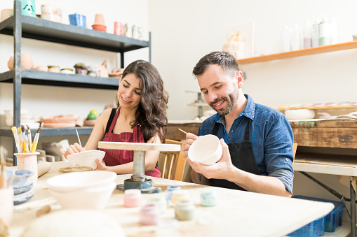 Smiling Couple Doing Creative Painting On Bowls In Pottery Workshop