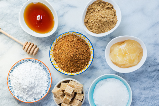 Different Kinds of Sugar and Sweeteners in the Bowls, such as coconut, pure cane, icing, maple syrup, dark brown soft sugar, honey, demerara cubes