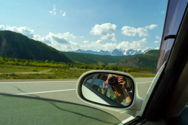 Photo of Reflection in the mirror of a riding white car of photographer who makes photo of a large altai mountains with snow on top by a sunny bright day with a blue clear sky green field and clouds