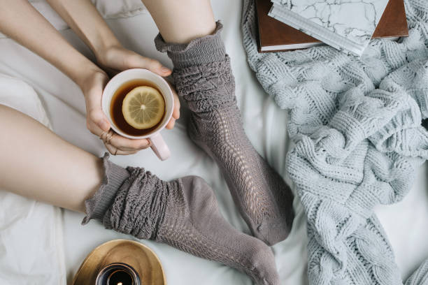 cozy flatlay of woman's legs in warm grey stockings in bed with knitted sweater and books holding cup of lemon tea, selective focus - stockings human leg female women imagens e fotografias de stock