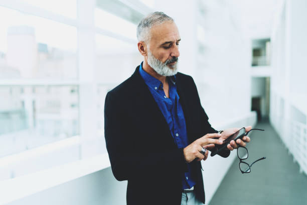 istock Aged bearded man wearing smart casual clothes typing messages on his mobile phone while standing in the light corridor in the building. CEO reading business emails on his smartphone. 1029975884