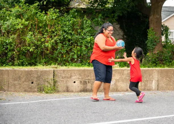 The body-positive, cheerful happy Latino, Mexican-American woman playing ball outdoor with her little daughter in the sunny hot summer day at the parking lot nearby his house in Pennsylvania, USA