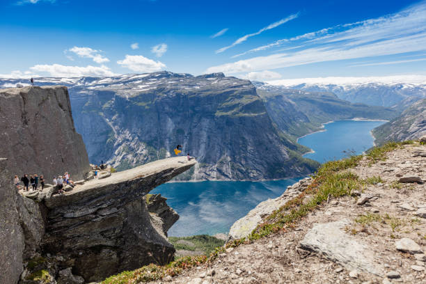A male hiker is making a somersault on Trolltunga in Norway. This is a main tourist attraction in the Odda Region in southern Norway. Lots of hikers in the back are standing in a queue to get an image of each other.