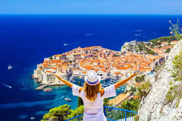 Happy young girl enjoys view of old town (medieval Ragusa) and Dalmatian Coast of Adriatic Sea in Dubrovnik. Blue sea with white yachts, beautiful landscape, aerial view, Dubrovnik, Croatia