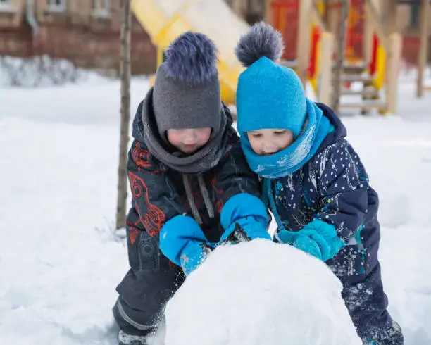 Two nice children boys 2 and 4 years in winter roll big snowball on playground