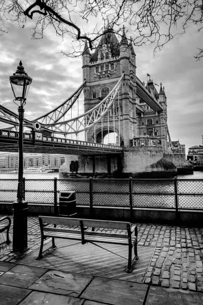 This is a black and white image of Tower Bridge in London.  It shows an empty bench which would be a good place to site and enjoy the view.