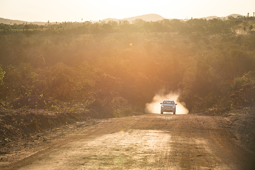 Silver SUV speeding uphill on a countryside road in Bonito, Mato Grosso do Sul, Brazil. Dust cloud behind the car as it drives on a dirt road in dry weather during sunset hour.