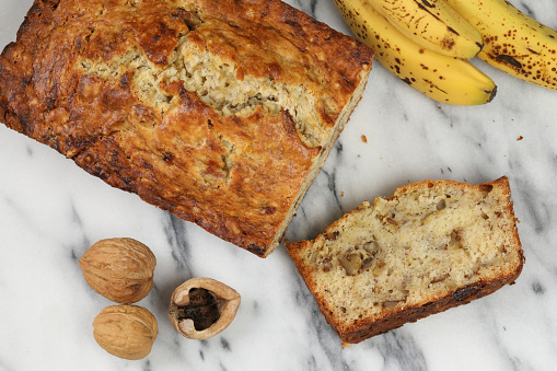 An overhead close up horizontal photograph of a loaf of banana bread, a slice of the cake, some fresh walnuts and several over ripened bananas.