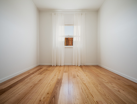 Wide angle interior shot of an empty room with a timber window, wood floors and curtains. View to outside is of a wooden fence.