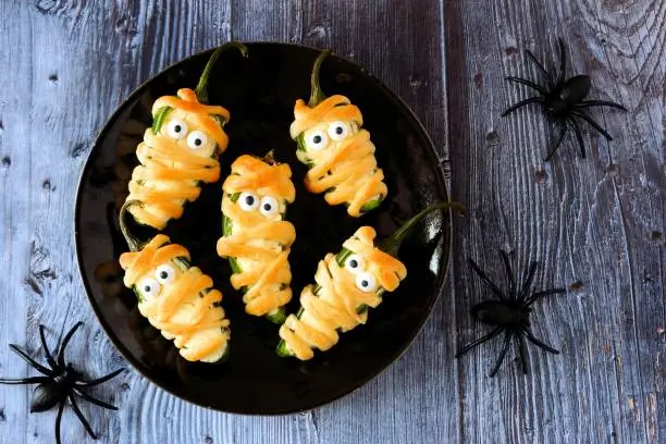 Plate of Halloween mummy jalapeno poppers, top view with a dark wood background with spiders