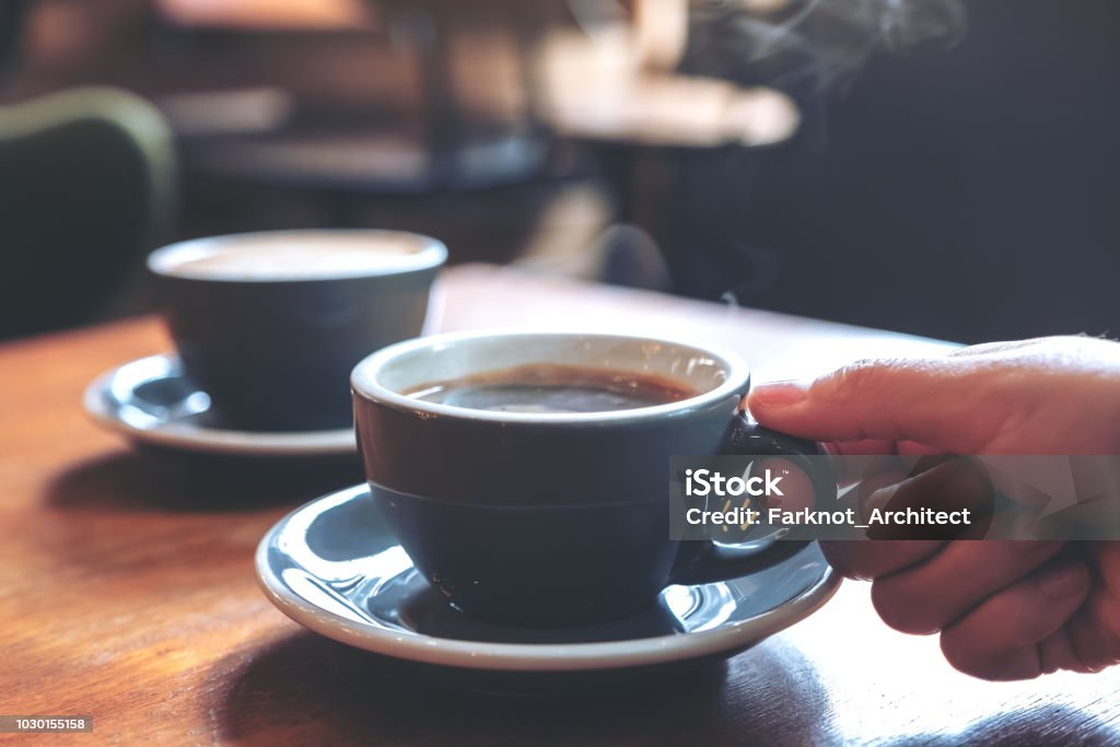 カフェの木製テーブルで煙でホット コーヒーのブルーのカップを持っている手のクローズ アップ画像 - カフェのロイヤリティフリーストックフォト