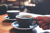 Closeup image of a hand holding a blue cup of hot coffee with smoke on wooden table in cafe
