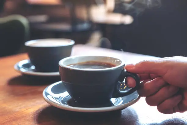 Photo of Closeup image of a hand holding a blue cup of hot coffee with smoke on wooden table in cafe