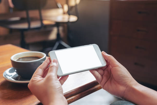 istock Mockup image of hands holding and using a white mobile phone with blank screen horizontally for watching with coffee cup on wooden table 1030155250