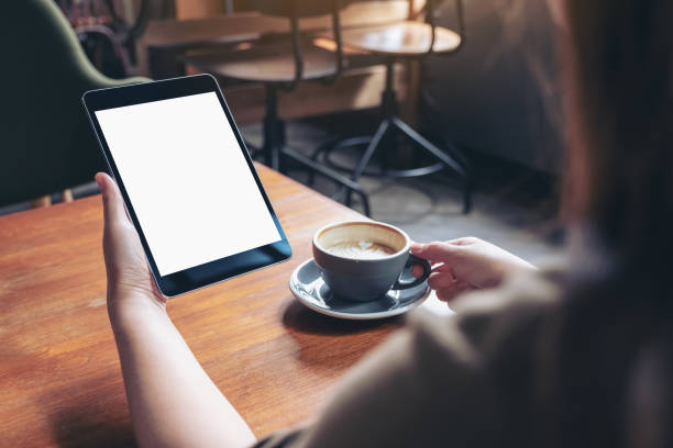 istock Mockup image of woman holding black tablet pc with blank white screen while drinking coffee on wooden table in cafe 1030155300