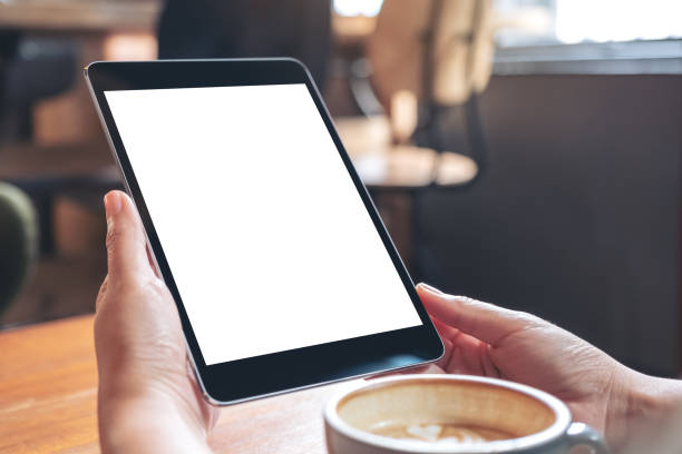 istock Mockup image of hands holding black tablet pc with blank white screen with coffee cup on wooden table in cafe 1030155308