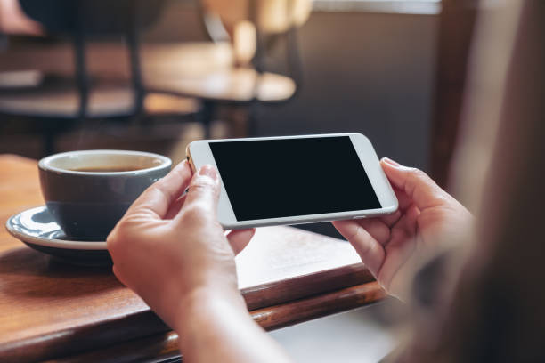 istock Mockup image of hands holding and using a white mobile phone with blank black screen horizontally for watching with coffee cup on wooden table 1030155352