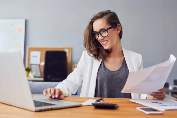 Smiling young woman working in home office using laptop and documents