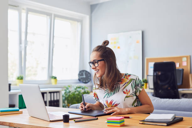 Cheerful attractive young female graphic designer smiling and working at her desk in modern office Cheerful attractive young female graphic designer smiling and working at her desk in modern office design professional stock pictures, royalty-free photos & images