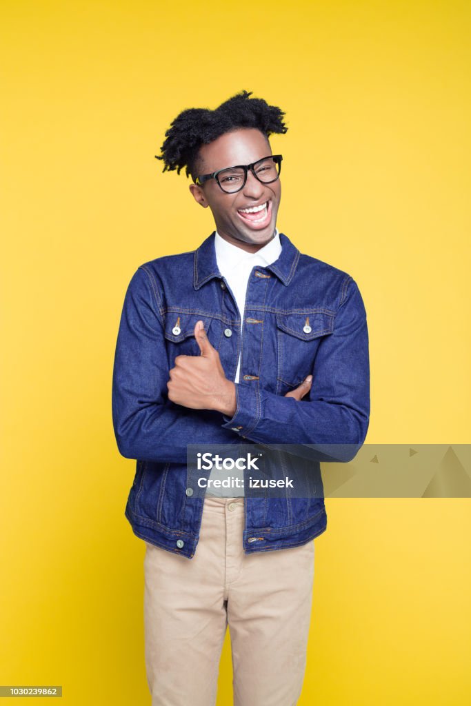 Funny 80's style portrait of cheesy nerdy young man Funny portrait of cheesy nerdy young afro american man wearing oversized jeans jacket and glasses, standing with arms crossed against yellow background. 1980-1989 Stock Photo