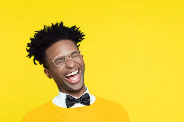 Funny portrait of happy nerdy young afro American man wearing yellow sweater and black bow tie laughing with eyes closed.