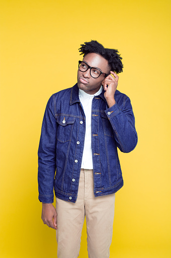 Portrait of nerdy young afro American man wearing oversized jeans jacket and glasses, standing against yellow background.