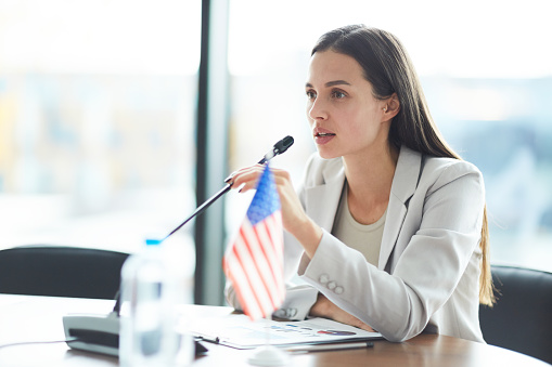 Portrait of confident young woman giving speech to microphone while sitting at table in conference room, copy space