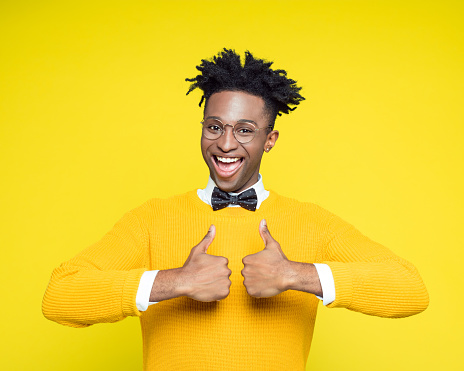 Funny portrait of happy nerdy young afro American man wearing yellow sweater and black bow tie laughing at the camera with thumbs up.