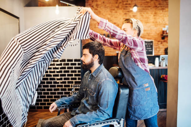 Young female barber spreading cape on customer. Hairdresser preparing client for shave. They are in barber shop.