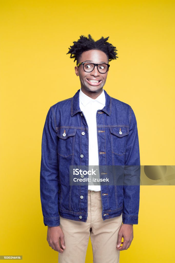 Funny 80's style portrait of cheesy nerdy young man Funny portrait of cheesy nerdy young afro american man wearing oversized jeans jacket and glasses, standing against yellow background. Cheesy Grin Stock Photo