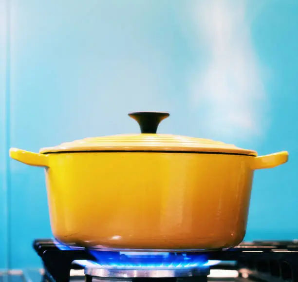 A yellow enamel casserole, with its lid on, sits on a lit gas burner creating steam as it heats.