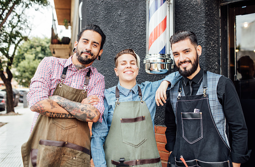 Portrait of smiling barbers standing outside hair salon. Multi-ethnic male and female hairdressers are wearing aprons. They are against barber's pole.