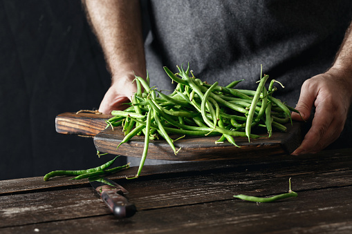 Male hands holding raw green bean on wooden cutting board on dark background. Healthy, vegetarian food concept