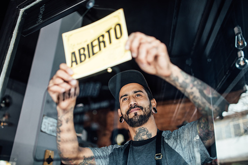 Low angle view of mid adult barber hanging an open sign on door. Male owner is holding abierto sign seen through glass. He is working in hair salon.