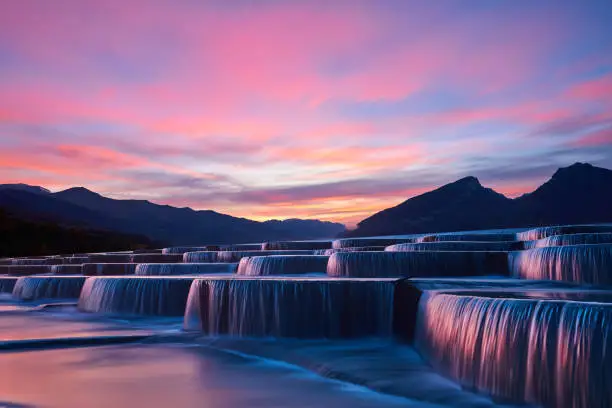 Side view of stepped waterfall group at sunrise in pink sky.