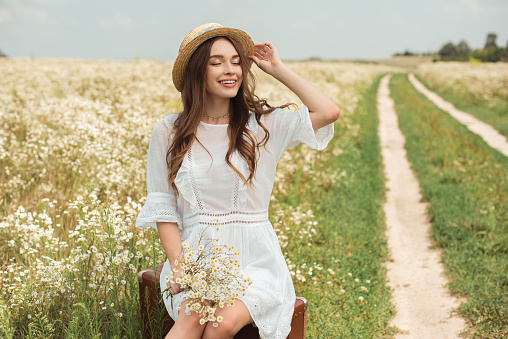 smiling woman in white dress with bouquet of wild camomile flowers sitting on retro suitcase in field