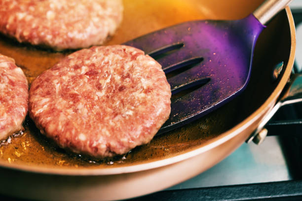 A spatula turns a raw hamburger patty in a frying pan.