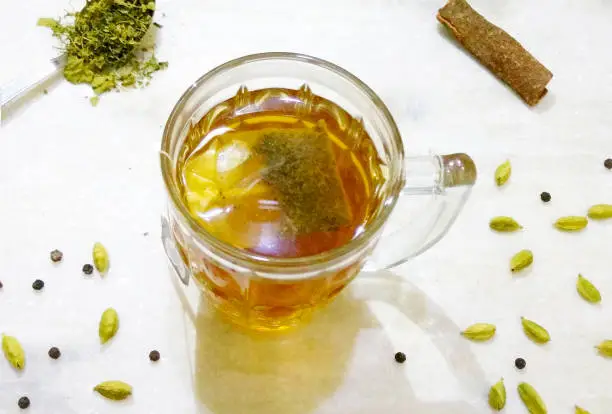 Mug of Organic Green Tea in Glass Cup made up of Honey & Tulsi with Cinnamon Stick, Cardamon and Black Pepper placed on white background